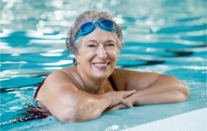 A senior in a swimming pool with her elbows up on the edge smiling, with blue goggles