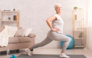 A senior woman in her living room doing a lunge with her right foot forward to stretch out her legs to remain limber