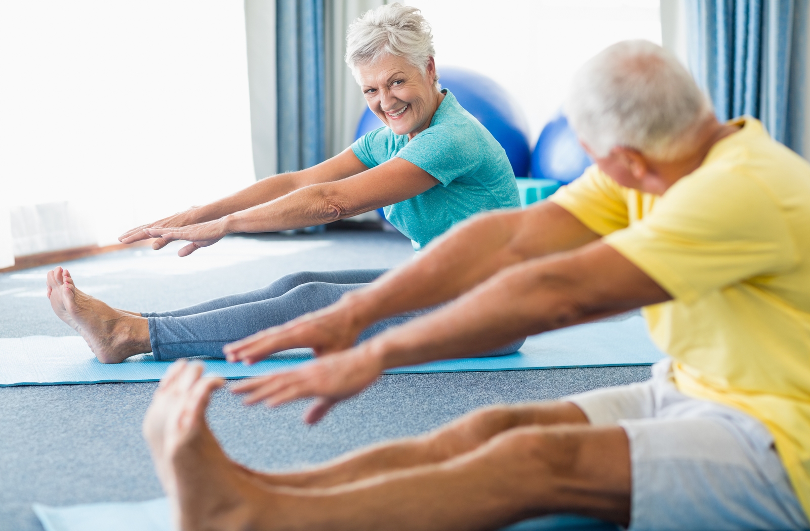 Two seniors sitting on yoga mat's reaching for their toes to stretch their muscles to improve their range of motion and flexibility