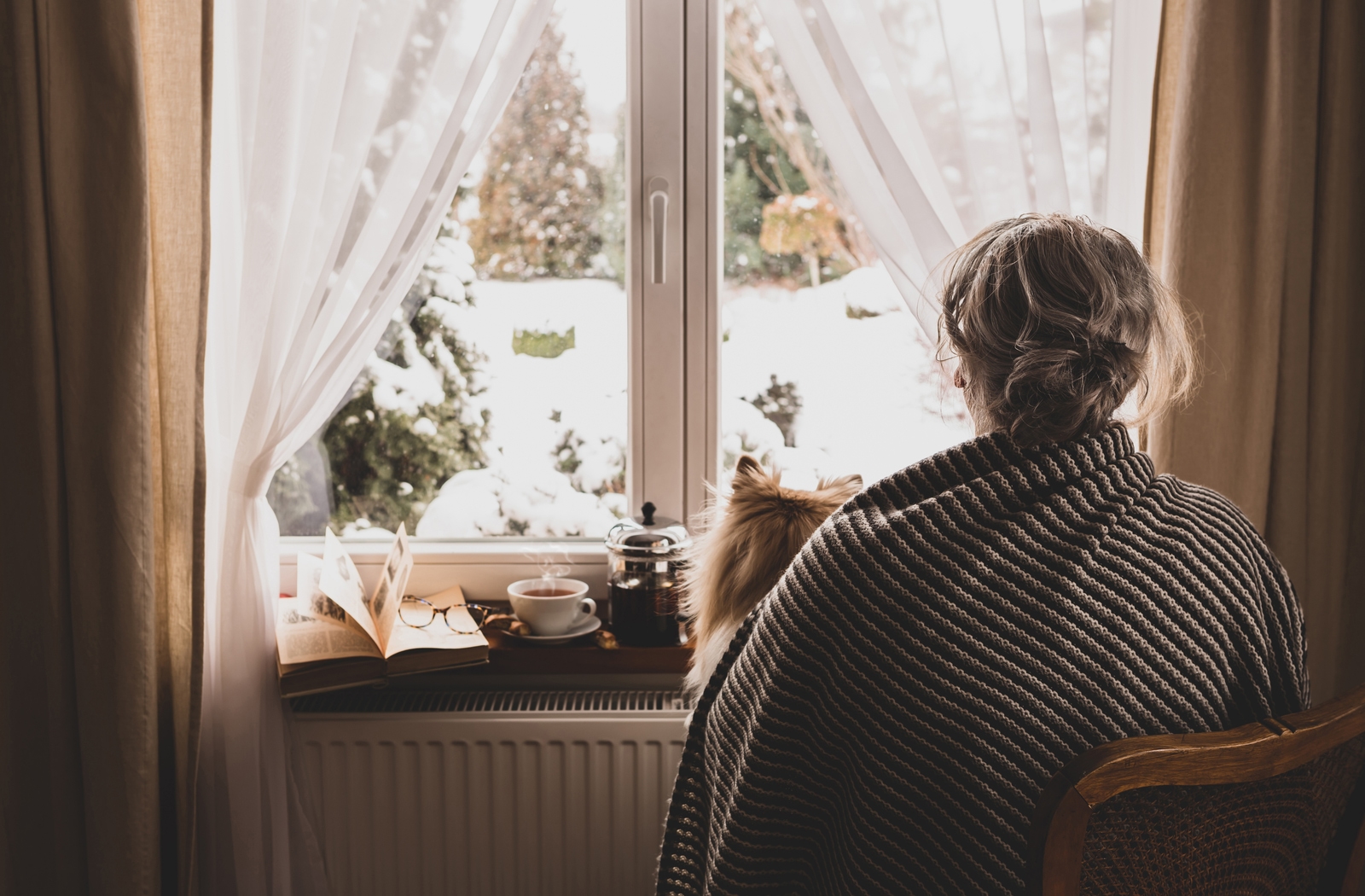 A view from behind a senior woman wrapped in a blanket, looking out the window during the winter time suffering from Seasonal Affective Disorder