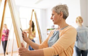 A senior woman holding a colored pencil drawing on a canvas, participating in an art class with other seniors