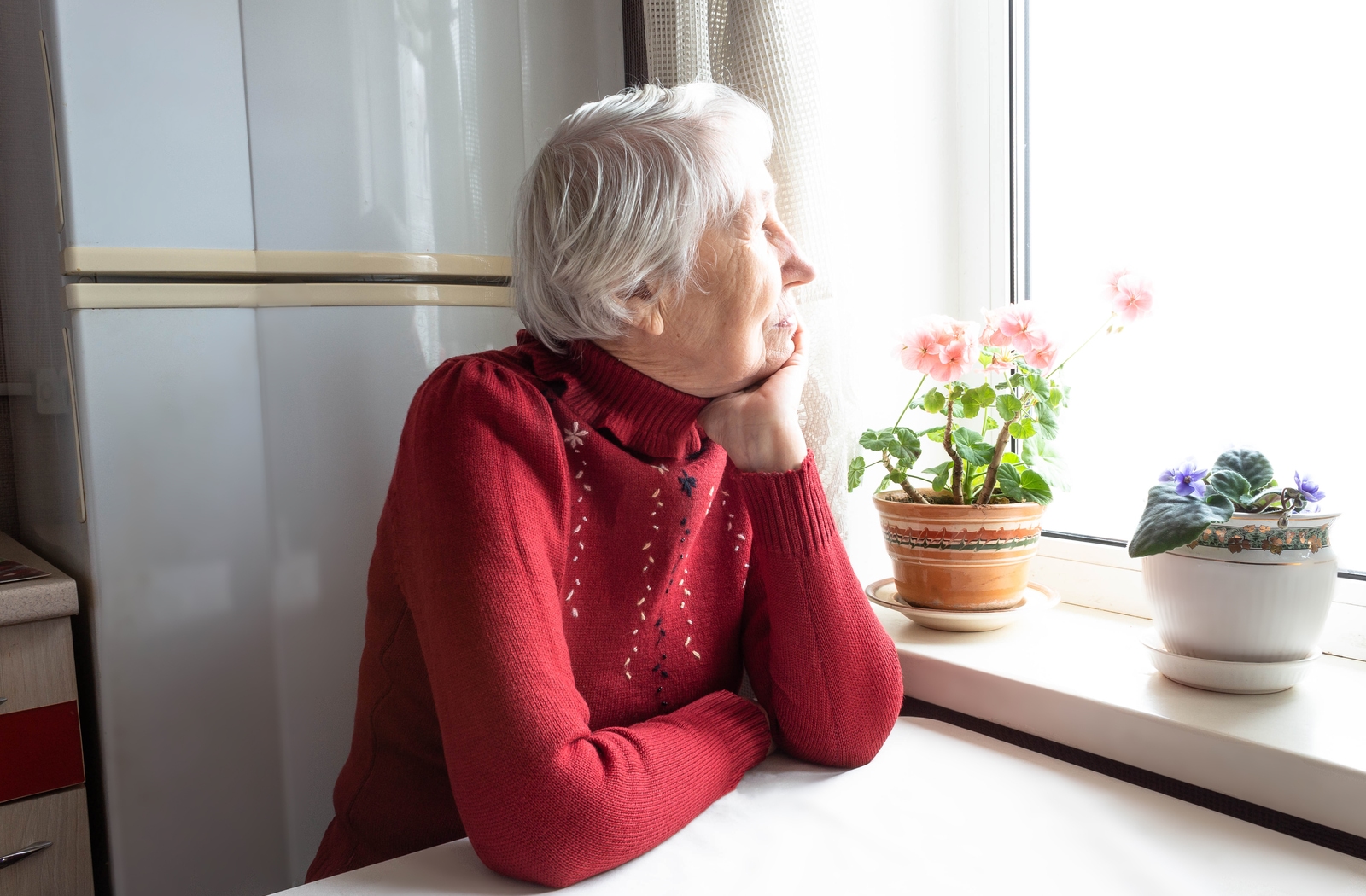 A senior woman wearing a red shirt sitting at a table in her kitchen, resting her chin on her arm looking out the window feeling a sense of loneliness