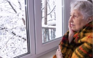 A senior woman looking out the window in the winter with a gloomy look on her face suffering from Seasonal Affective Disorder