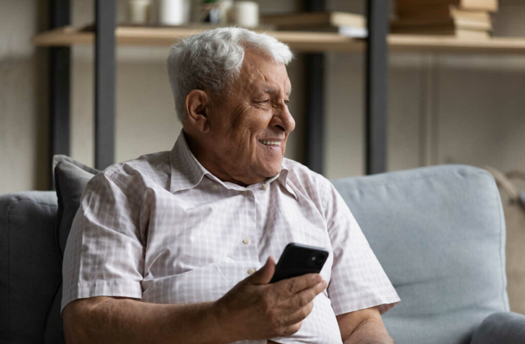 A senior man with gray hair smiling and sitting on a couch while holding a smartphone.