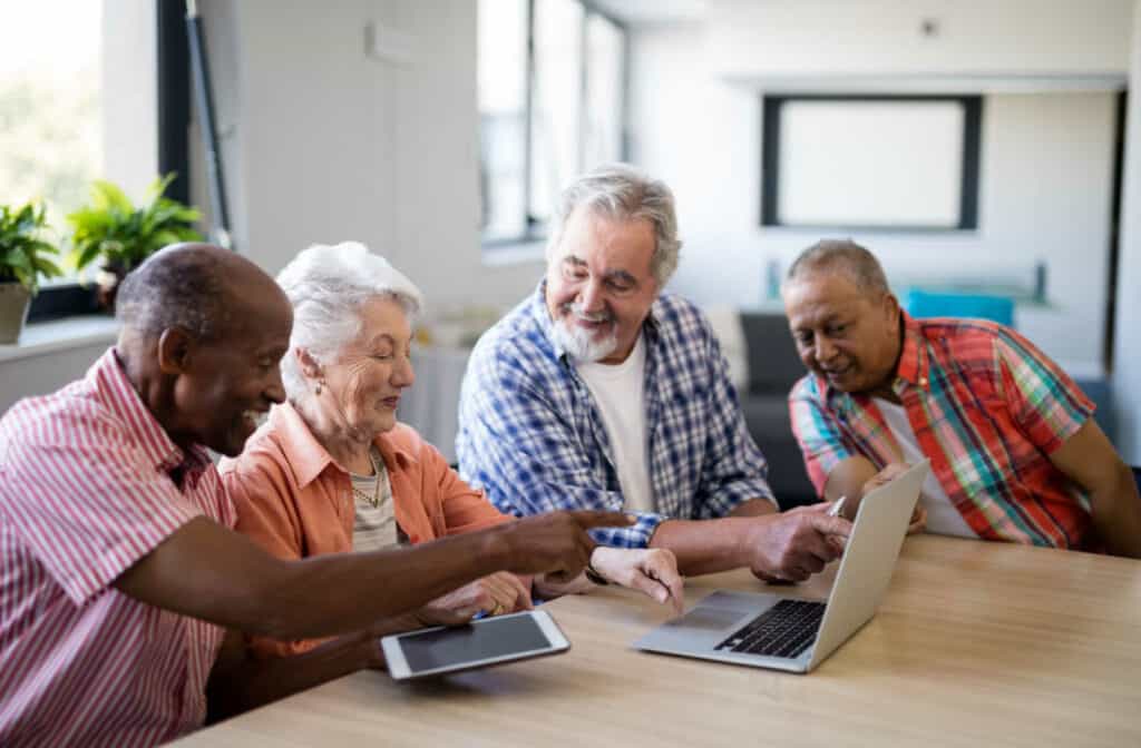 A group of seniors smiling and talking to each other while using a laptop and a tablet.