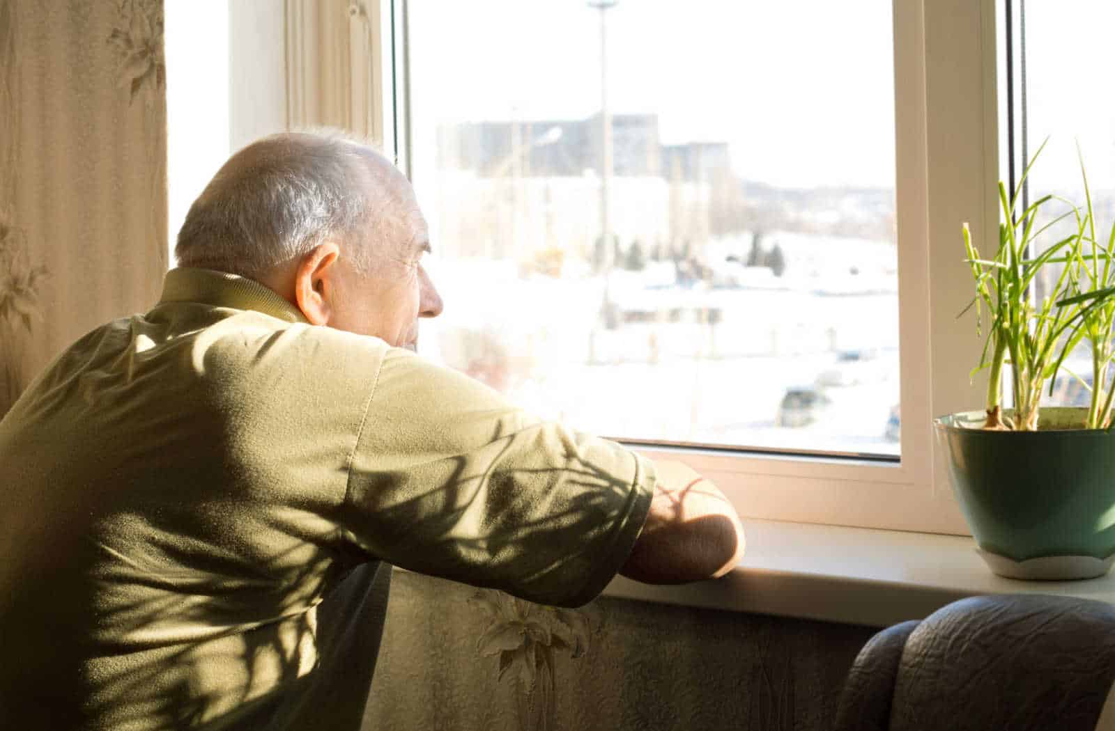 A senior man is staring out of a window, feeling lonely and isolated.