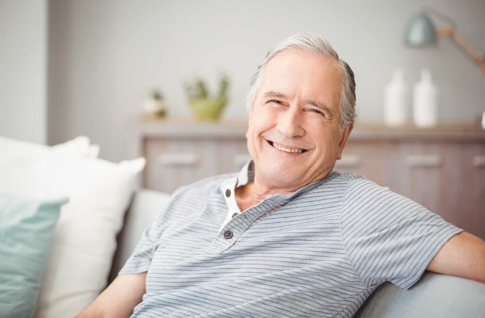 A senior man sitting on a couch, smiling and looking directly at the camera